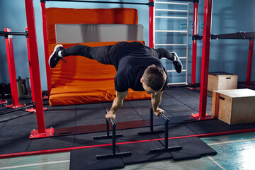 Muscular, athletic man in sportswear training in gym indoors, doing Straddle Planche exercises with parallel bars. Concept of active and healthy lifestyle, body care, fitness, sport
