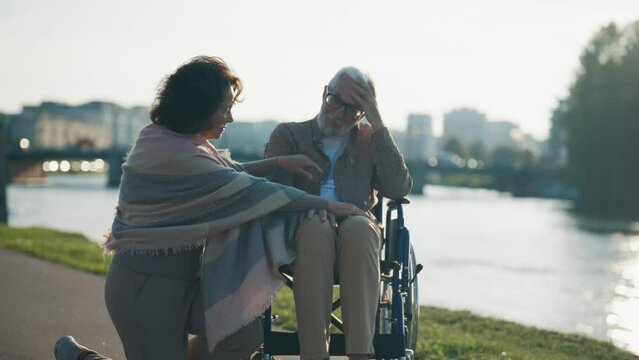 Wife helps husband to sit in wheelchair. Woman putting senior man legs on wheelchair footplates, walking together in city park. Support male help male with special needs, physical disability concept.