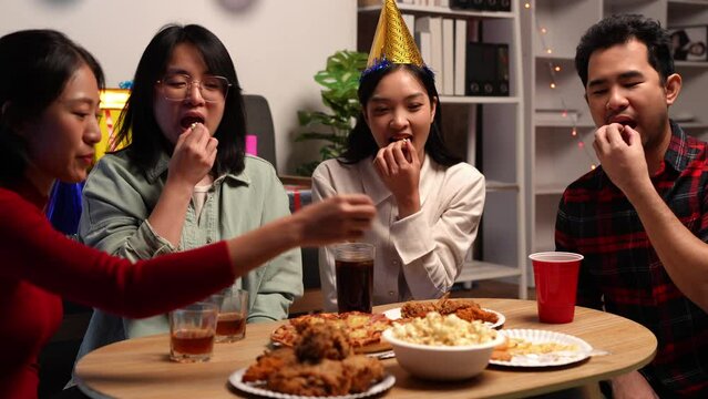 Group of happy young friends celebrating at a party with snacks popcorn pizza and drinks. The atmosphere at the event was full of laughter while sitting at the dining table together in the home office