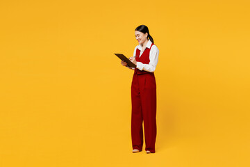 Full body young lawyer employee business woman of Asian ethnicity she wearing red vest shirt work at office hold clipboard paper account documents isolated on plain yellow background. Career concept.