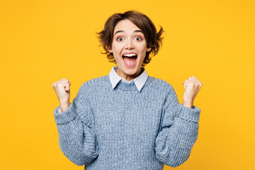 Young overjoyed happy woman wears grey knitted sweater shirt casual clothes doing winner gesture celebrate clench fists say yes isolated on plain yellow background studio portrait. Lifestyle concept.