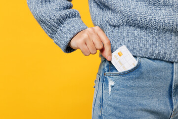 Close up cropped young woman she wears grey knitted sweater shirt casual clothes hold in hand put mock up of credit bank card into pocket isolated on plain yellow background studio. Lifestyle concept.