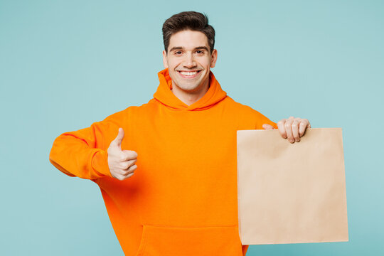 Young man wears orange hoody casual clothes hold brown clear blank craft paper takeaway bag mock up show thumb up isolated on plain pastel light blue cyan color background studio. Lifestyle concept
