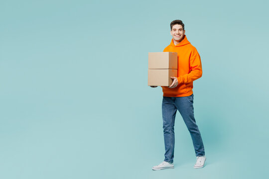 Full body smiling happy fun young man wears orange hoody casual clothes hold stack cardboard blank boxes isolated on plain pastel light blue cyan color background studio portrait. Lifestyle concept