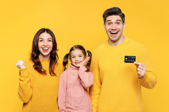 Young happy parents mom dad with child kid girl 7-8 years old wear pink knitted sweater casual clothes hold credit bank card do winner gesture isolated on plain yellow background. Family day concept.