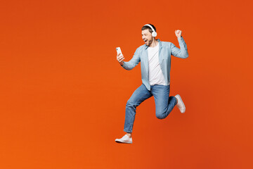 Full body young man wears blue shirt white t-shirt casual clothes jump high hold in hand use mobile cell phone listen to music in headphones isolated on plain red orange background. Lifestyle concept. - obrazy, fototapety, plakaty