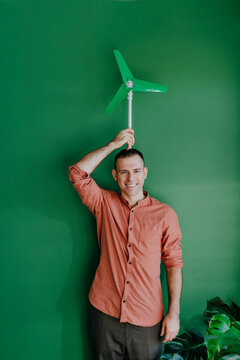 Happy freelancer holding wind turbine model on head in front of green wall