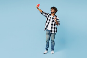 Full body young Indian man wear shirt white t-shirt casual clothes doing selfie shot on mobile cell phone post photo on social network show v-sign isolated on plain blue background. Lifestyle concept - obrazy, fototapety, plakaty
