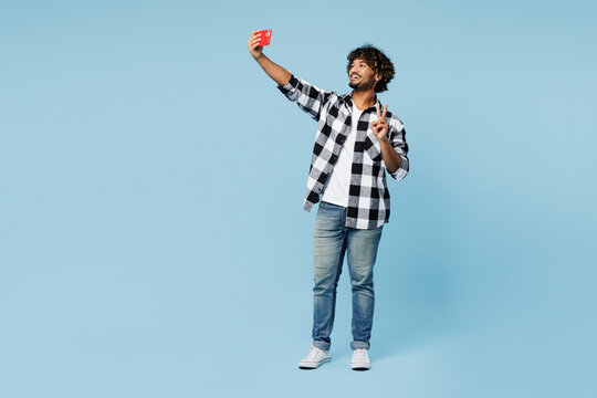 Full body young Indian man wear shirt white t-shirt casual clothes doing selfie shot on mobile cell phone post photo on social network show v-sign isolated on plain blue background. Lifestyle concept