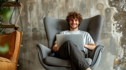 Young Caucasian man with curly hair, comfortably seated in a modern gray armchair, smiling and relaxed while using a laptop on his lap