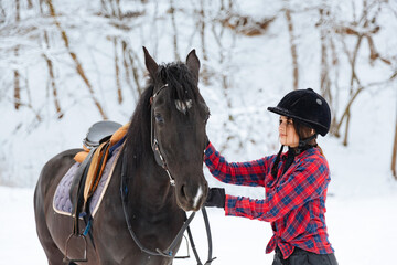 A beautiful brunette girl in a plaid black-and-white shirt walks with a big black horse in a snowy...