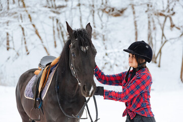 A beautiful brunette girl in a plaid black-and-white shirt walks with a big black horse in a snowy park. A woman in a helmet for riding walks in a snowy forest with a thoroughbred horse