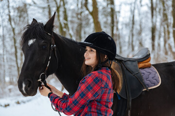 A beautiful brunette girl in a plaid black-and-white shirt walks with a big black horse in a snowy...