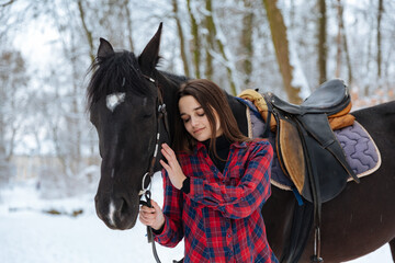 A beautiful brunette girl in a plaid black-and-white shirt walks with a big black horse in a snowy park. A woman in a helmet for riding walks in a snowy forest with a thoroughbred horse