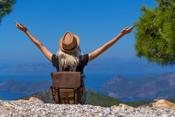 Rear view of an enthusiastic woman in a hat with a backpack with her hands raised, looking from a height at an amazingly beautiful landscape of mountains and the sea. Concept of freedom and adventure.