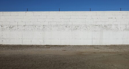 Long white concrete block wall with barbed wire on top. Asphalt street in front, blue sky above. Background for copy space. 
