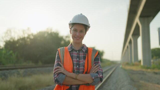 Beautiful engineer woman in safety uniform and helmet standing with arms crossed on railroad smiling and looking at camera. Engineer leadership concept