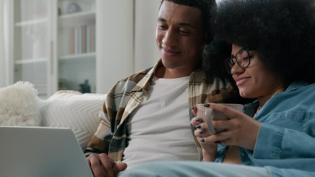 African American couple at home couch looking at laptop talking watching film movie discuss choosing goods online store internet shopping order delivery using computer together woman drinking tea cup