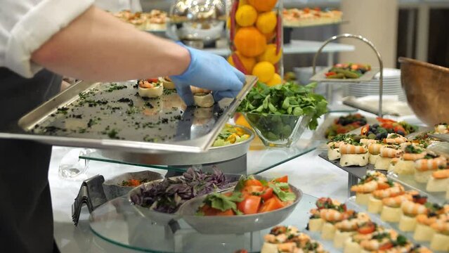 Close-up of waiter's hands in blue gloves lay out canapes with shrimps, cherry tomatoes and herbs on the buffet table in a restaurant. Buffet food in a restaurant.