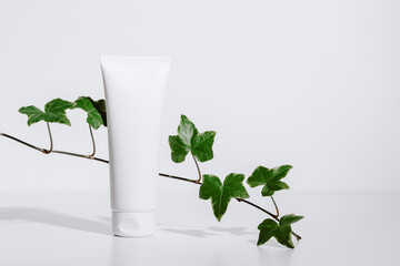 White mockup cosmetic tube for facial cream with green plant leaves and shadows on white...