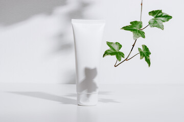 White mockup cosmetic tube for facial cream or lotion with green plant leaves and shadows on white...