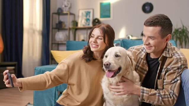 Satisfied Caucasian couple taking family photo with their lovely dog in room. People sitting on floor and hugging happy dog. Young wife holding phone while husband petting labrador retriever.