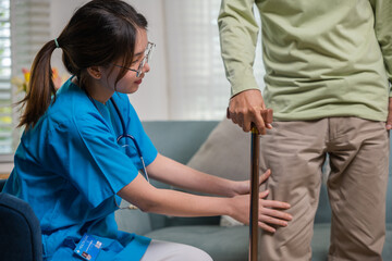 Asian young woman nurse checking knee and leg after surgery of senior old man patient suffering from pain in knee, doctor asking elderly man about pain symptom with walking stick