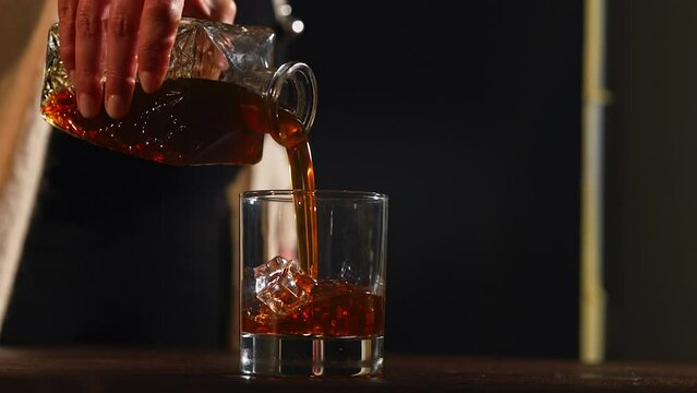 Close up of a barlady  pouring whiskey in a cold glass full of ice cubes. Pouring thick liquor into an old fashion glass. Cocktail made with bourbon on a wooden table a dark background.