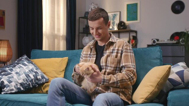 Handsome Caucasian man holding his furry cat while sitting on blue couch in living room. Happy man petting cute red kitty. Lovely domestic pets at home. Resting with his little friend.