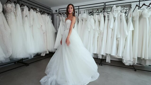 A beautiful young bride tries on a white wedding dress against the background of a large number of dresses in the store. Having fun and spinning.