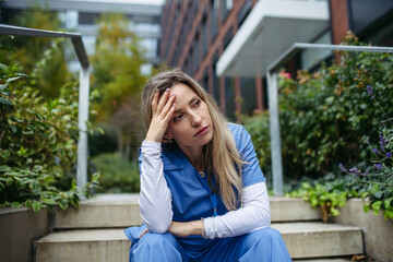 Young female doctor feeling overwhelmed at work, sitting on stairs, looking at phone. Healthcare...