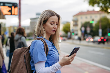 Female nurse going home from work, waiting for bus, scrolling on smartphone. Work-life balance of...