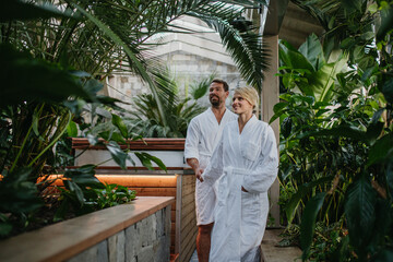 Beautiful couple standing by hot tub, wearing bathrobes, enjoying romantic wellness weekend in spa....