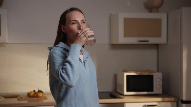Beautiful brunette woman in a blue sweater drinks water standing in the kitchen. A thirsty woman takes a sip of pure mineral water from a glass.
