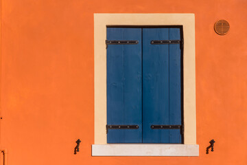 Una finestra di legno dipinta di blu sul muro arancione di una casa del borgo di Caorle piccola località di mare in provincia di Venezia