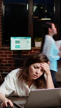 Vertical video Businesswoman working on company project overnight, feeling frustrated, putting head in hands. Upset employee sighing while looking over figures on laptop screen, exasperated by failure