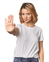 Fototapeta premium Blonde middle-aged Caucasian woman in studio standing with outstretched hand showing stop sign, preventing you.