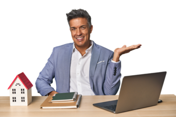 Middle-aged Hispanic businessman with real estate theme showing a copy space on a palm and holding another hand on waist.