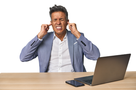 Elegant businessman at desk with laptop covering ears with fingers, stressed and desperate by a loudly ambient.