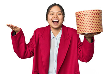 Young Chinese woman holding a heart-filled box receiving a pleasant surprise, excited and raising...