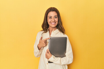Middle-aged woman with laptop on yellow smiling and pointing aside, showing something at blank...