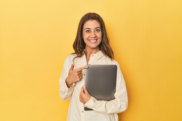 Middle-aged woman with laptop on yellow smiling and pointing aside, showing something at blank...