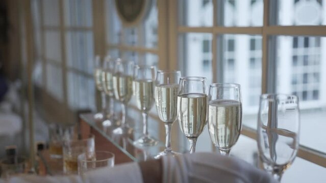 A human hand in a white glove is holding a clear glass with liquid. There are multiple glasses with golden-colored liquid in the background, likely champagne.