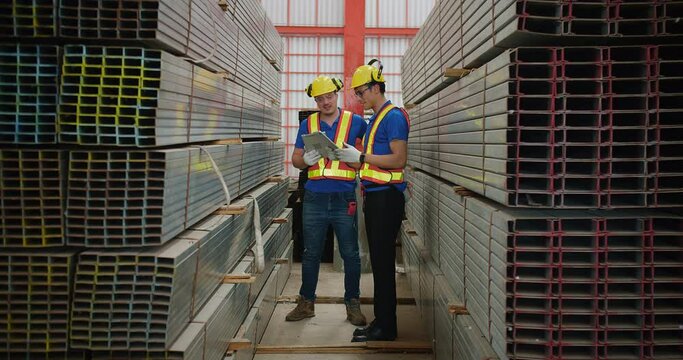 Two construction workers in hard hats and reflective vests are having a discussion in a warehouse full of metal beams