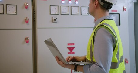 An engineer in a high-visibility vest and hard hat inspects systems on his laptop at an industrial control panel