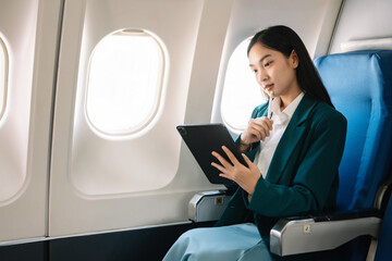 Fototapeta premium Attractive Asian female passenger of airplane sitting in comfortable seat while working laptop and tablet with mock up area using wireless connection.