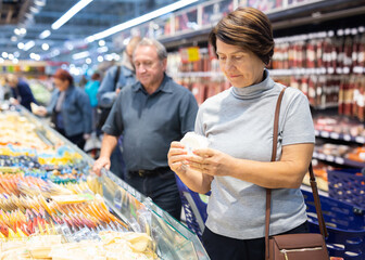 Mature woman diligently choosing fresh cheese in supermarket