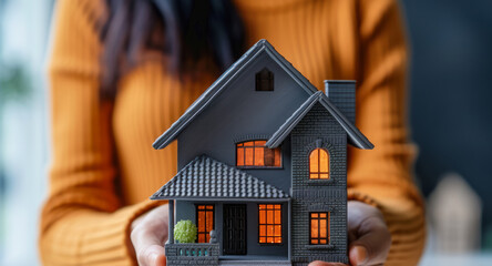 Close-up of a real estate agent's female hands holding a dark-colored model of a small house in her palm. Concept of buying a house, real estate, construction business - obrazy, fototapety, plakaty