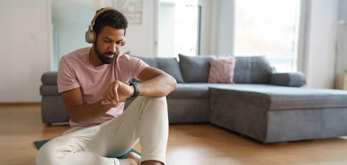 Young man resting after home workout, checking his exercise performance on smartwatch. Morning or evening workout routine, listening to music through wireless headphones.