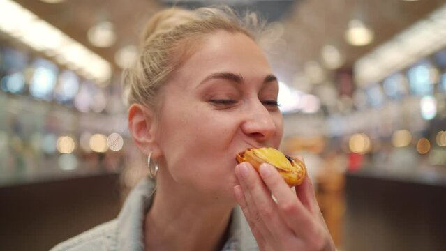 Beautiful young female eating Portuguese dessert in market. Happy tourist woman enjoying traditional dessert Pastel de Nata in city. Blond lady on vacation.